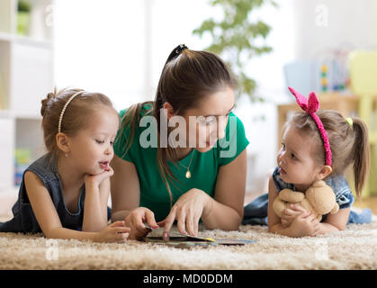 Giovane madre la lettura di un libro per i suoi figli delle figlie. I bambini e la mamma che giace sul tappeto nel soggiorno accogliente. Foto Stock