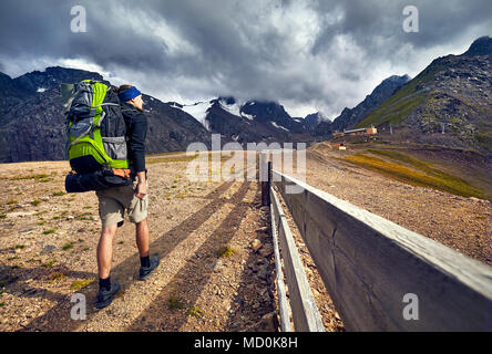 Escursionista in con grande zaino verde a piedi sulle montagne innevate a nuvoloso scuro dello sfondo del cielo in Shymbulak Ski Resort in Kazakistan Foto Stock