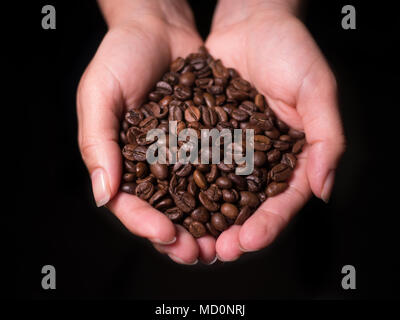 Mani femminili holding freschi chicchi di caffè tostati closeup da sopra a coppa con scuri su sfondo nero Foto Stock