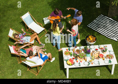 Felice giovani agghiacciante, parlando e suonando la chitarra in campagna grigliata, amici seduta a prendere il sole sulle sedie a sdraio Foto Stock