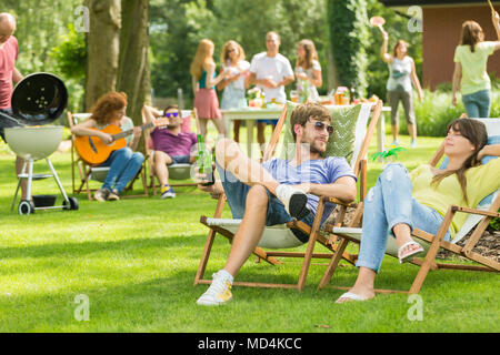 Giovani amici avente picnic barbecue nella natura, suonare la chitarra, giocare a badminton, godendo della soleggiata giornata estiva all'aperto Foto Stock