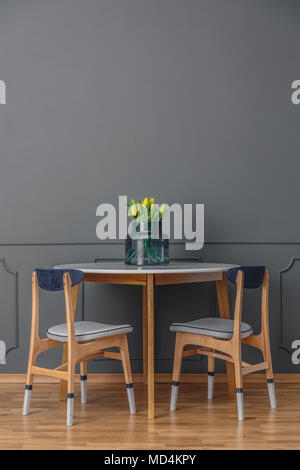 Vernice-immerso sedie e una semplice tavola di legno con un bouquet di tulipani gialli nel vaso di vetro da un vuoto, muro grigio in uno stile minimalista sala da pranzo interi Foto Stock