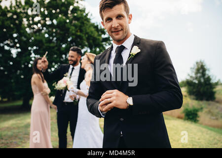 Ritratto di bello groom permanente al parco con la sua sposa e amici prendendo selfie in background. Lo sposo dopo la cerimonia nuziale. Foto Stock