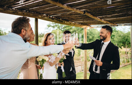 Valutazione la tostatura con una bottiglia di champagne e coppie in viaggio di nozze. I giovani sposi bicchieri tintinnanti e godendo il momento con ospite al ricevimento di nozze al di fuori. Foto Stock