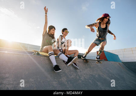 Due giovani donne seduta sul pattinaggio rampa con una ragazza di praticare lo skateboard. Donna di skateboard skateboard di equitazione a skate park rampa con gli amici si Foto Stock