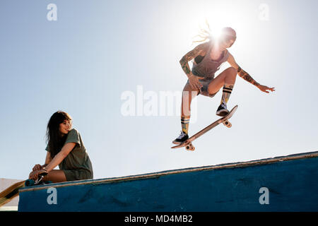 Skater femmina corse su skateboard con femmina amico seduto sulla rampa. Per la corsa su skateboard femmina a skate park con un amico a guardare la routine. Foto Stock