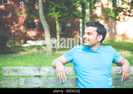 Closeup ritratto, sorridenti, regolari Giovane Uomo in camicia blu seduti sulla panca in legno, rilassati guardando al lato, isolare gli alberi di sfondo, boschi. Ri Foto Stock