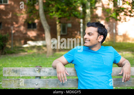 Closeup ritratto, sorridenti, regolari Giovane Uomo in camicia blu seduti sulla panca in legno, rilassati guardando al lato, isolare gli alberi di sfondo, boschi Foto Stock