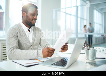 Vita-up ritratto di allegro African American businessman studiare il documento mentre è seduto alla scrivania di modern open plan office, lens flare Foto Stock