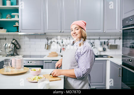 Giovane bella femmina cuocere in grembiule e hat rendendo insalata di frutta in cucina moderna e sorridente in telecamera felicemente Foto Stock