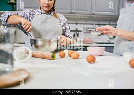 Basso angolo vista del giovane cuoca e il suo collega irriconoscibile sbattere le uova e setacciare la farina attraverso un setaccio in cucina Foto Stock