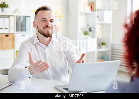Giovane bello barbuto office manager in camicia bianca seduta a tavola con laptop, chiacchierando con un collega e sorridente Foto Stock