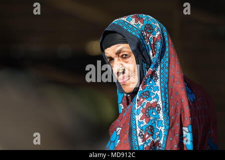 Nizwa, Oman - 23 Mar 2018: Vecchia Omani donna in piedi da solo al sole con un hijba ed espressione seria mentre strizzare gli occhi con gli occhi su un backgroun scuro Foto Stock