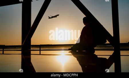 Silhouette di un turista guy guardando il decollo del piano seduta alla finestra di airport al tramonto di sera. Concetto di viaggio, la gente in aeroporto. Foto Stock