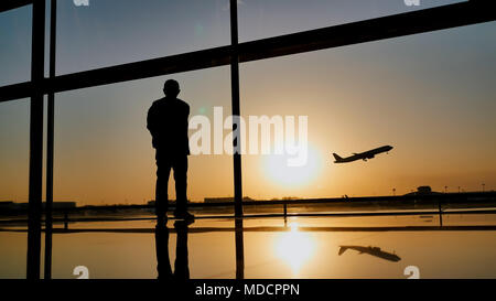 Silhouette di un turista guy guardando il decollo del piano in piedi alla finestra di airport al tramonto di sera. Concetto di viaggio, la gente in aeroporto. Foto Stock