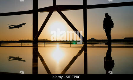 Silhouette di un turista guy guardando il decollo del piano in piedi alla finestra di airport al tramonto di sera. Concetto di viaggio, la gente in aeroporto. Foto Stock