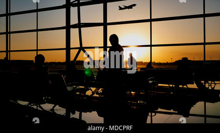 Silhouette di un turista guy guardando il decollo del piano in piedi alla finestra di airport al tramonto di sera. Concetto di viaggio, la gente in aeroporto. Foto Stock