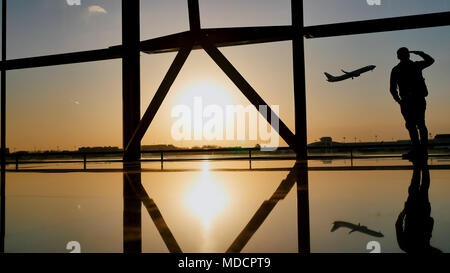 Silhouette di un turista guy guardando il decollo del piano in piedi alla finestra di airport al tramonto di sera. Concetto di viaggio, la gente in aeroporto. Foto Stock