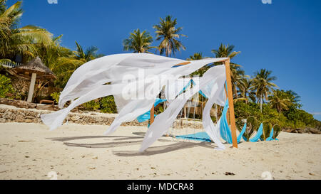 Arco di nozze decorata con fiori sulla spiaggia vicino all'oceano. Il suo bianco e blu tele volare nel vento. Cuori decorativi dalle foglie di palme. Sabbiosa costa tropicale. Foto Stock
