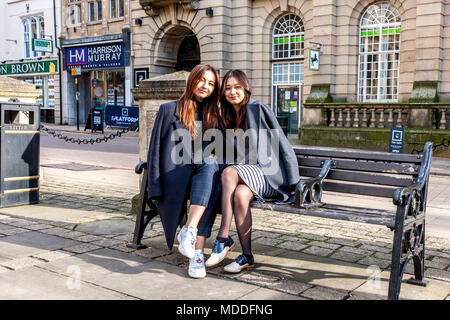 Due donne orientali amici da Northampton college seduti su un ferro battuto e panca in legno nella parte anteriore della Chiesa di Tutti i Santi, Northampton town ce Foto Stock