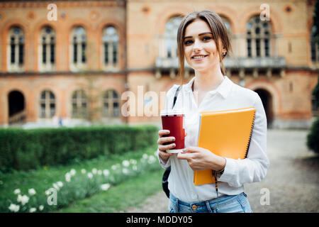 Sorridenti studente ragazza con zaino in università dello sfondo. Donna con le cartelle di lavoro e prendere la via di un caffè con il resto nel campus durante la pausa pranzo. Foto Stock