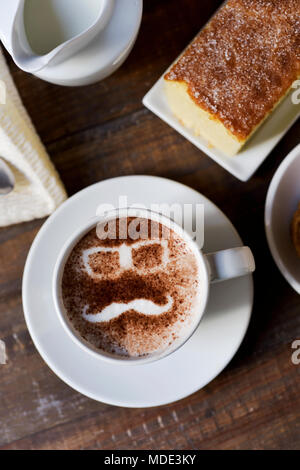 Una tazza di cappuccino con la silhouette di un paio di occhiali e un baffi formando il volto di un uomo disegnata con il cacao in polvere sulla sua schiuma, su un ta Foto Stock