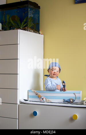 Carino il toddler ragazzo seduto in una grande barca di legno a casa, giocando con i pesci in legno, sorridente Foto Stock