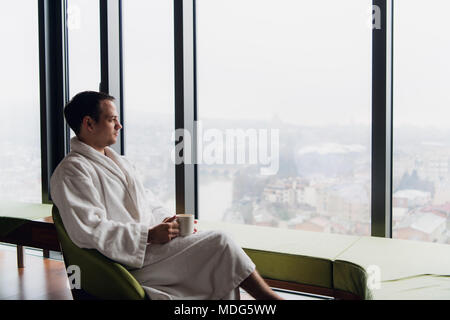 Giovane uomo godendo la sera caffè e tramonto stupendo panorama della città mentre in piedi dalla finestra Foto Stock