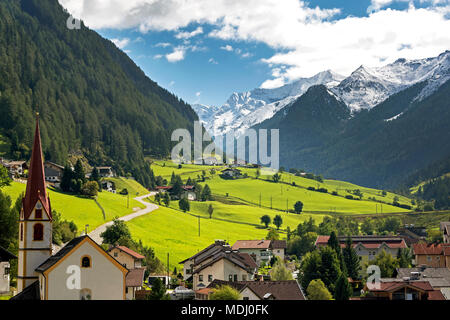 Villaggio alpino con campanile, prati verdi nella valle e montagne coperte di neve in lontananza; St. Jodok, Tirolo, Austria Foto Stock