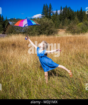 Una giovane ragazza sta in un campo di erba con ombrello alta e in equilibrio su un piede; Salmon Arm, British Columbia, Canada Foto Stock