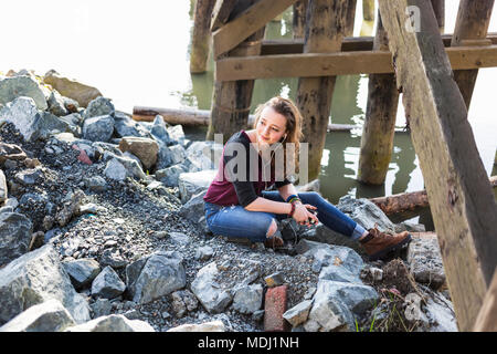 Ritratto di una giovane donna seduta sulle rocce sotto un ponte da acqua ascolto di musica Foto Stock