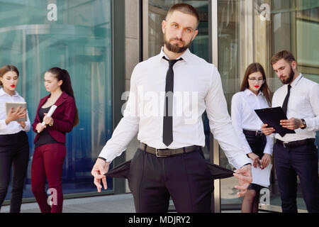 In bancarotta. Un giovane uomo in pantaloni neri e una camicia bianca dimostra la sua vuota tasche in background di un gruppo di impiegati di ufficio. Il licenziamento fro Foto Stock