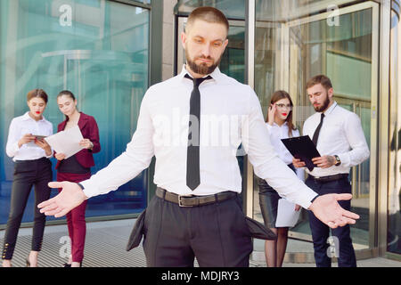 In bancarotta. Un giovane uomo in pantaloni neri e una camicia bianca dimostra la sua vuota tasche in background di un gruppo di impiegati di ufficio. Il licenziamento fro Foto Stock