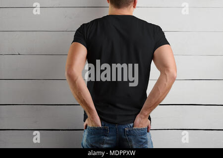 Il modello di un t-shirt nera su un uomo forte tiene le mani in tasca posteriore di jeans su uno sfondo di legno. Vista dal retro. Foto Stock