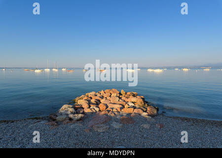 Massi accatastati sulla spiaggia al litorale del Lago di Garda Lago di Garda) con barche ancorate nella distanza, Bardolino in Veneto, Italia Foto Stock