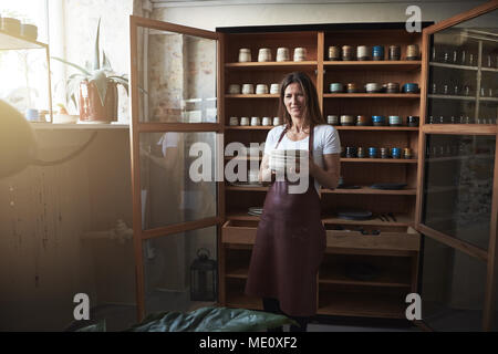 Donna sorridente artigiano in piedi nella parte anteriore del display ripiani nel suo studio di ceramiche azienda nuove piastre in ceramica Foto Stock