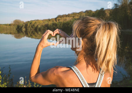 Giovane donna dalla Lakeshore facendo una forma di cuore dito telaio sul bellissimo paesaggio, la riflessione sulla superficie dell'acqua. Persone Viaggi amore ambiente Foto Stock