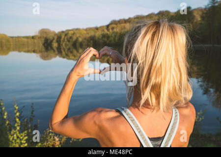 Giovane donna dalla Lakeshore facendo una forma di cuore dito telaio sul bellissimo paesaggio, la riflessione sulla superficie dell'acqua. Persone Viaggi amore ambiente Foto Stock