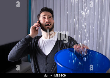 Preoccupato per il giovane uomo chiamando l'idraulico mentre acqua di perdita di cadere nella benna a casa Foto Stock