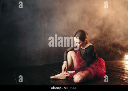 Ballerina in abito rosso seduta sul piano del palcoscenico del teatro e la legatura pointe scarpe. Eleganza ballerina la formazione in classe Foto Stock