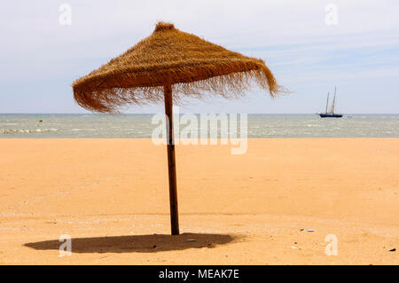 Ombrellone di paglia su una spiaggia di sabbia con una barca vela passato Foto Stock