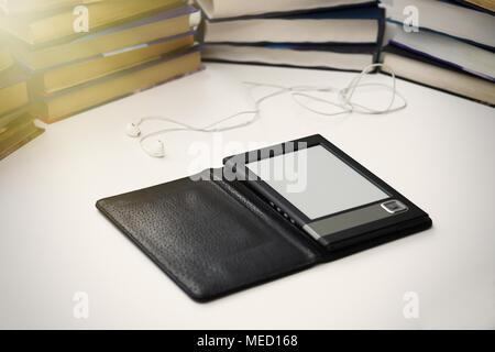 E-book si trova su un tavolo bianco su uno sfondo di libri di carta. Le cuffie si trovano nelle vicinanze. Foto Stock