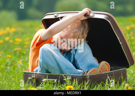 Grazioso piccolo bambino divertente cercando di nascondere all'interno di marrone vintage valigia. Ragazzo ride e sorride felicemente giocando all'aperto sul verde prato pieno Foto Stock