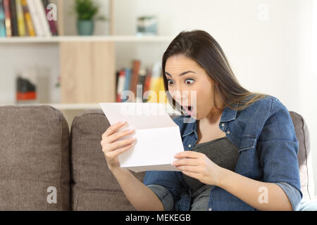 Stupiti donna lettura sorprendente notizia in una lettera seduta su un divano nel salotto di casa Foto Stock