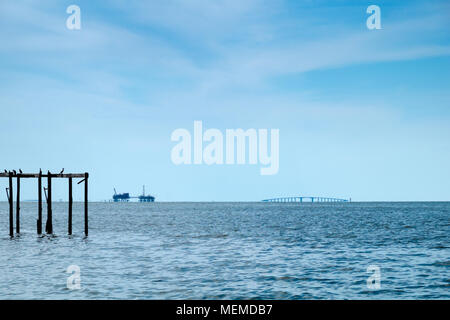 Nel tardo pomeriggio sulla Baia di Mobile, Alabama. Il gas naturale trivelle e Le Dauphin Island bridge sedersi all'orizzonte al di sotto di un azzurro intenso del cielo. Cormorani appollaiarsi su Foto Stock