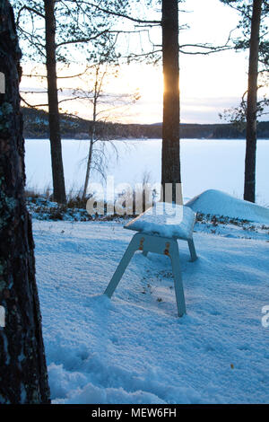 Una panca in legno è in piedi sulla riva di un coperto di ghiaccio sul lago. Foto Stock