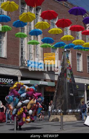 Venditore di palloncino in Exeter High Street, con la Exeter Riddle Obelisco Scultura e coloratissima primavera ombrelloni. Devon, Regno Unito. Aprile, 2018. Foto Stock