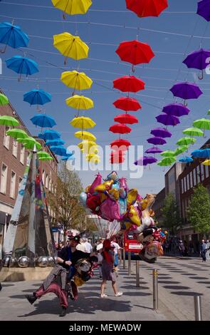 Venditore di palloncino in Exeter High Street, con la Exeter Riddle Obelisco Scultura e coloratissima primavera ombrelloni. Devon, Regno Unito. Aprile, 2018. Foto Stock