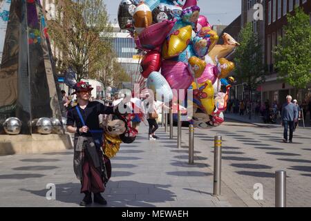 Venditore di palloncino in Exeter High Street, con la Exeter Riddle Obelisco Scultura e coloratissima primavera ombrelloni. Devon, Regno Unito. Aprile, 2018. Foto Stock