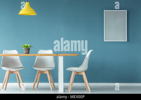 Sedie bianche al tavolo in legno con impianto sotto lampada gialla in blu sala da pranzo interno con poster Foto Stock